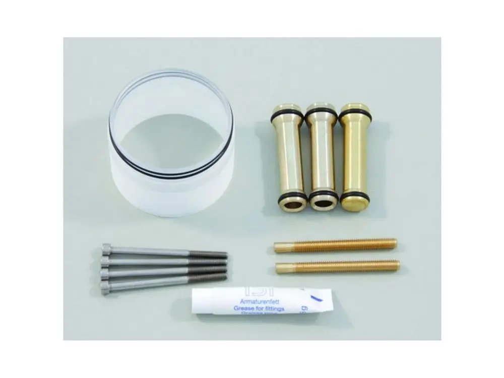 Ideal Standard Bauteile: Ein transparenter Zylinder mit O-Ringen, drei metallische Teile, zwei Schrauben und ein Tubus mit Schmiermittel auf einem flachen Untergrund.